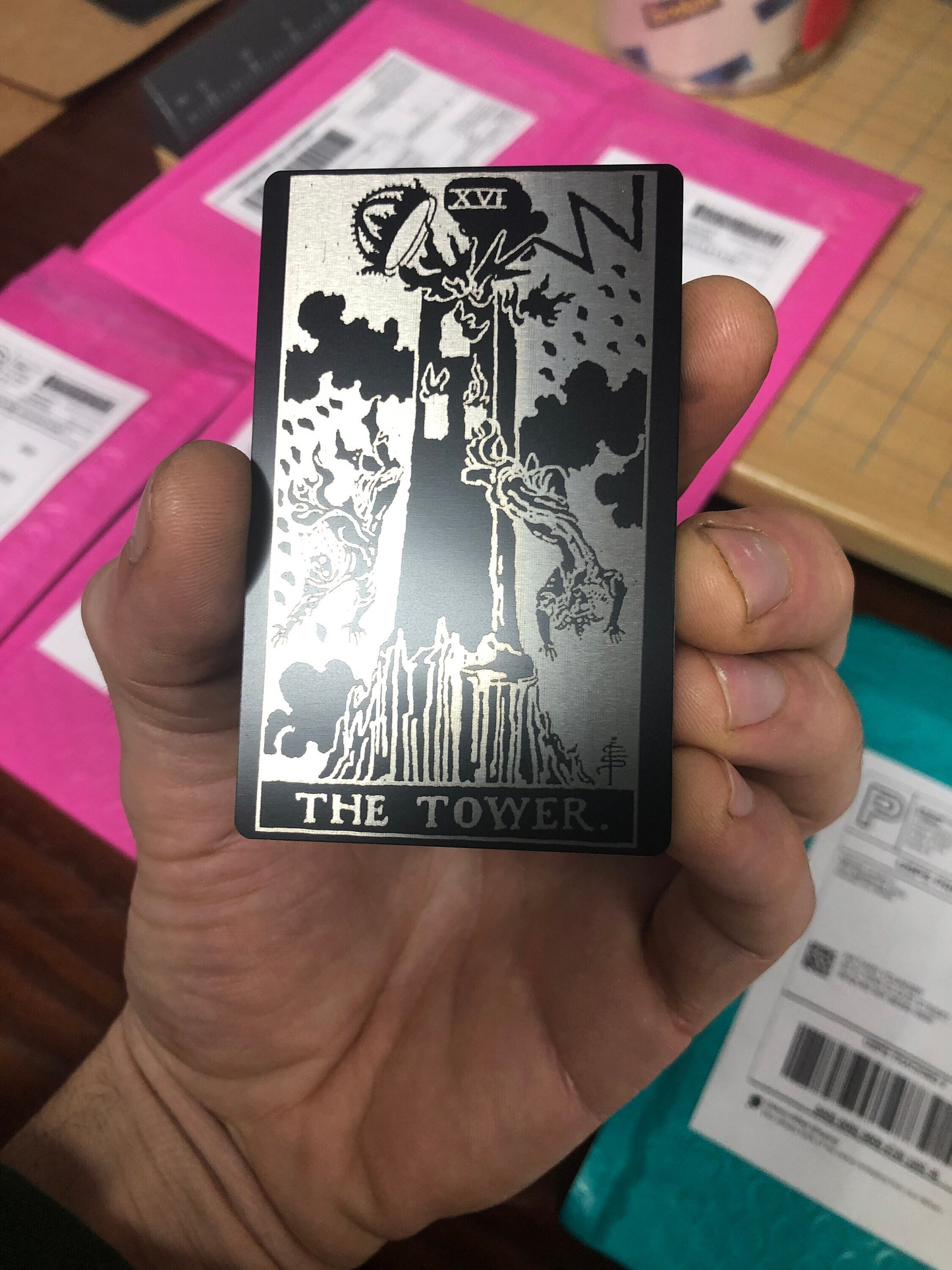 Hand holding a metal black/silver tarot card titled 'The Tower' with a black and white illustration.