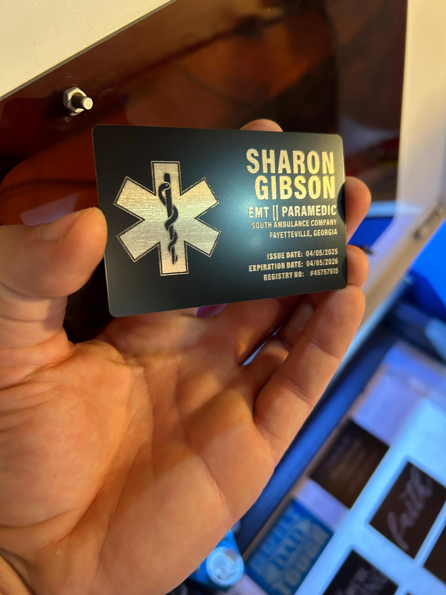 Hand holding a metal business card with personalized text example 'Sharon Gibson' and an EMT symbol.