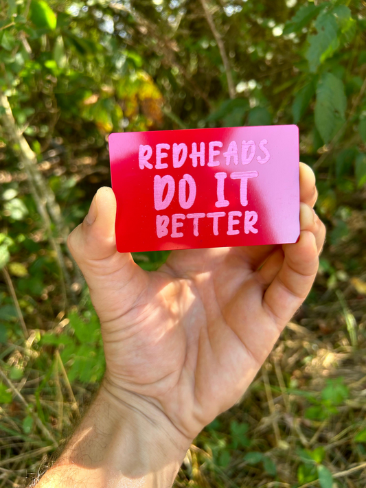 Hand holding a red card with 'Redheads Do It Better' text against a blurred natural background
