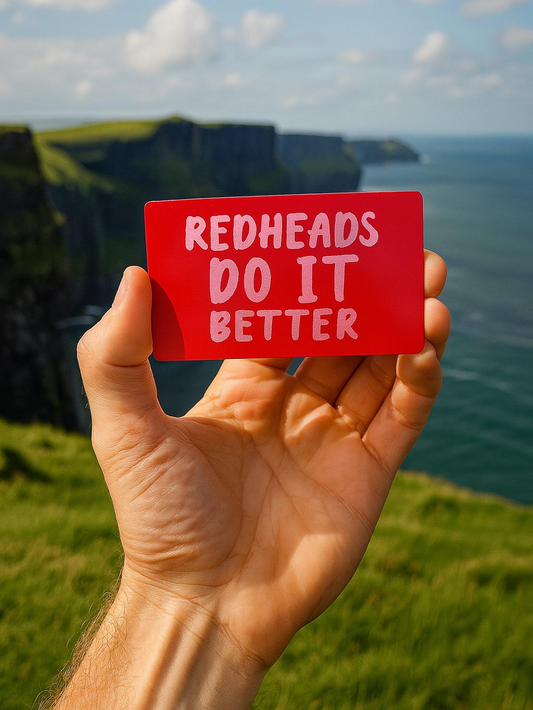 Hand holding a red card with 'REDHEADS DO IT BETTER' text against a scenic background.