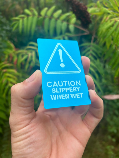 Hand holding a blue/silver metal warning sign with 'Caution Slippery When Wet' text against a green leafy background.