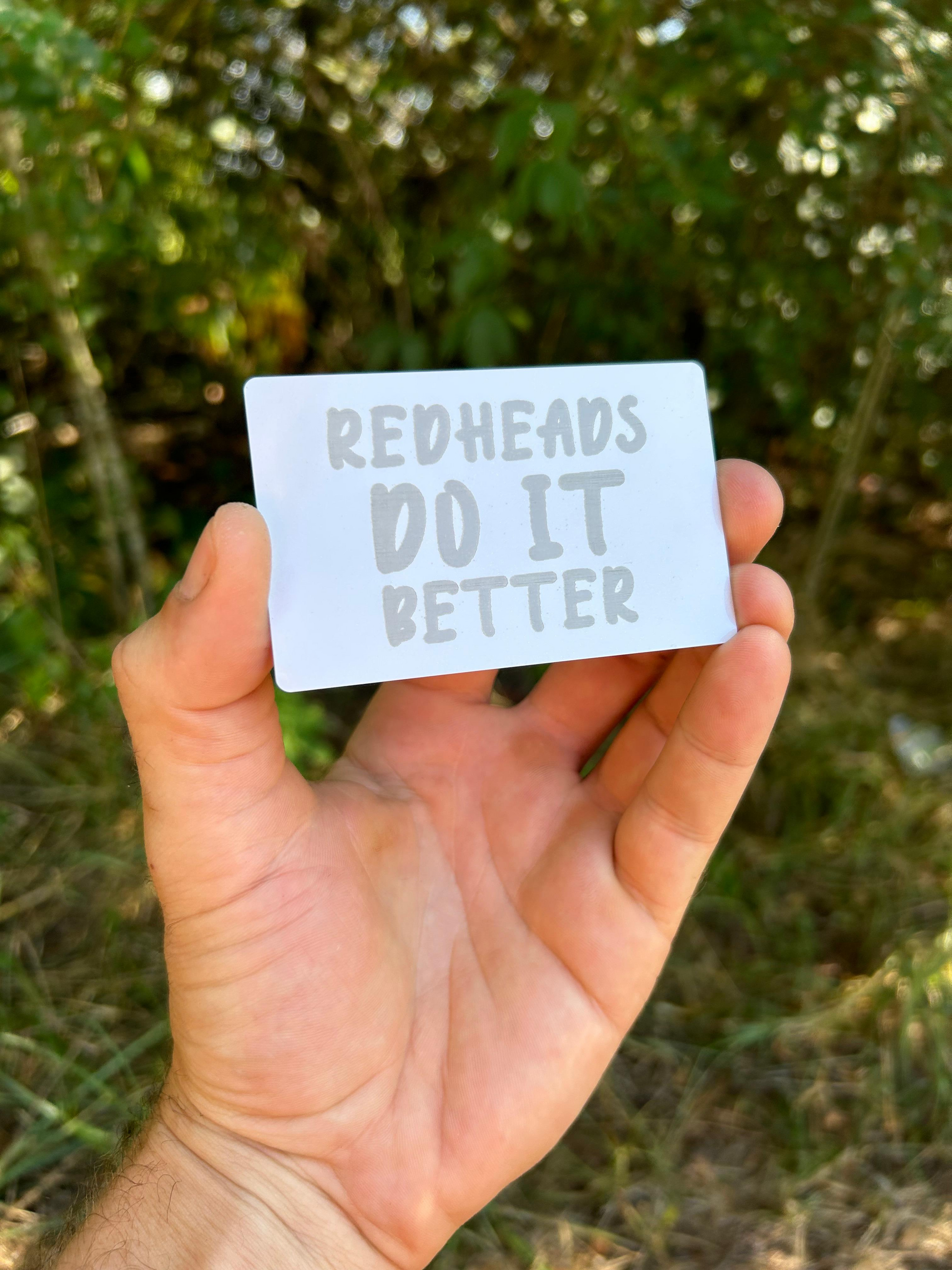 Hand holding a card with 'Redheads Do It Better' text against a blurred natural background