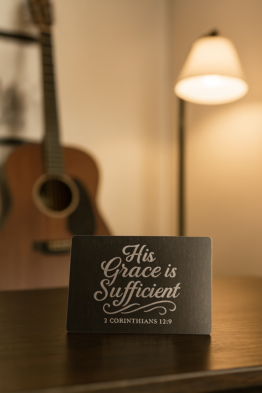 Decorative mini metal placardwith 'His Grace is Sufficient' text and 2 corinthians 12:9 bible verse reference on a wooden surface with a guitar in the background.