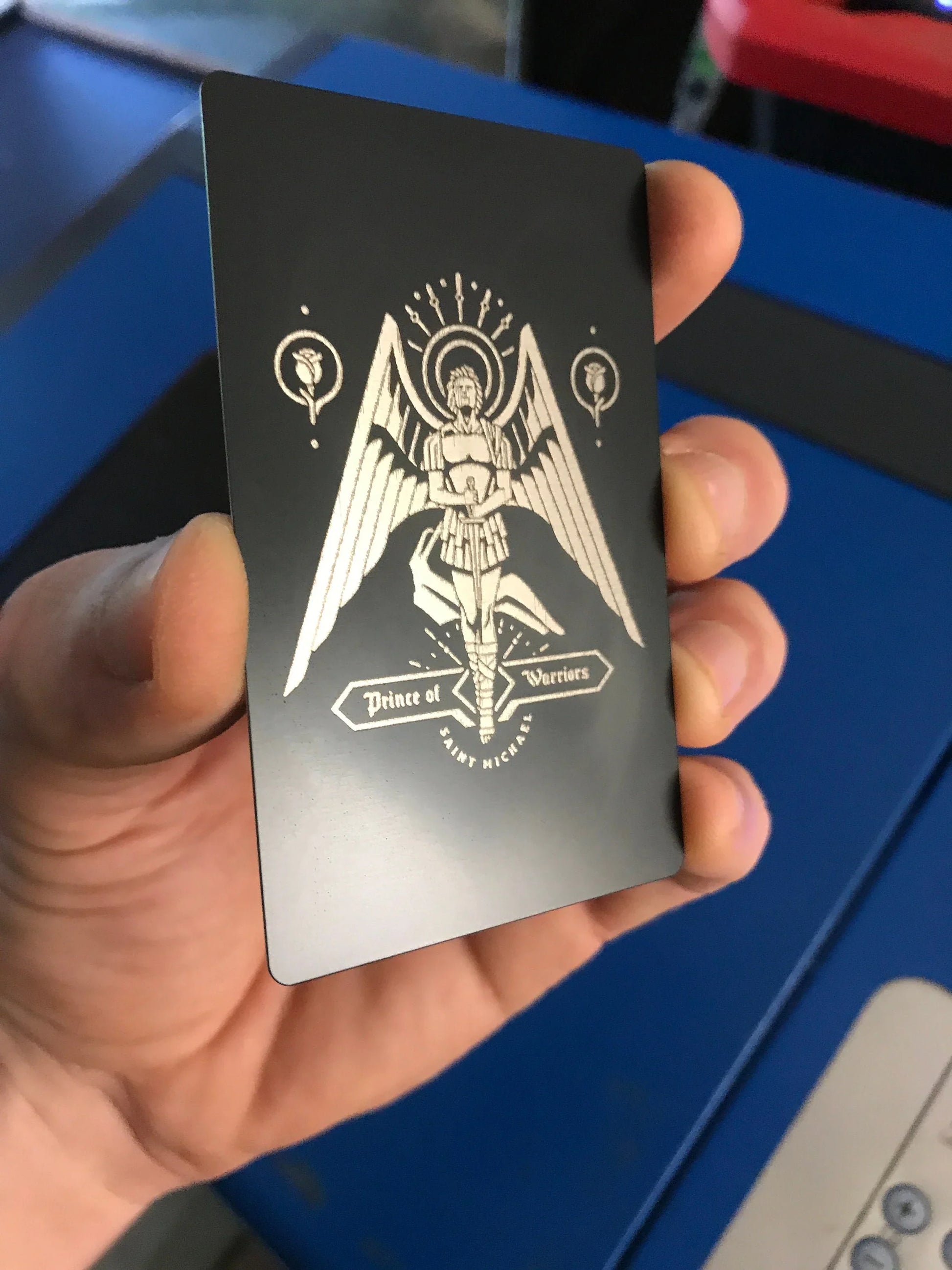 Hand holding a black card with a white St Michael Archangel design against a blue machine background