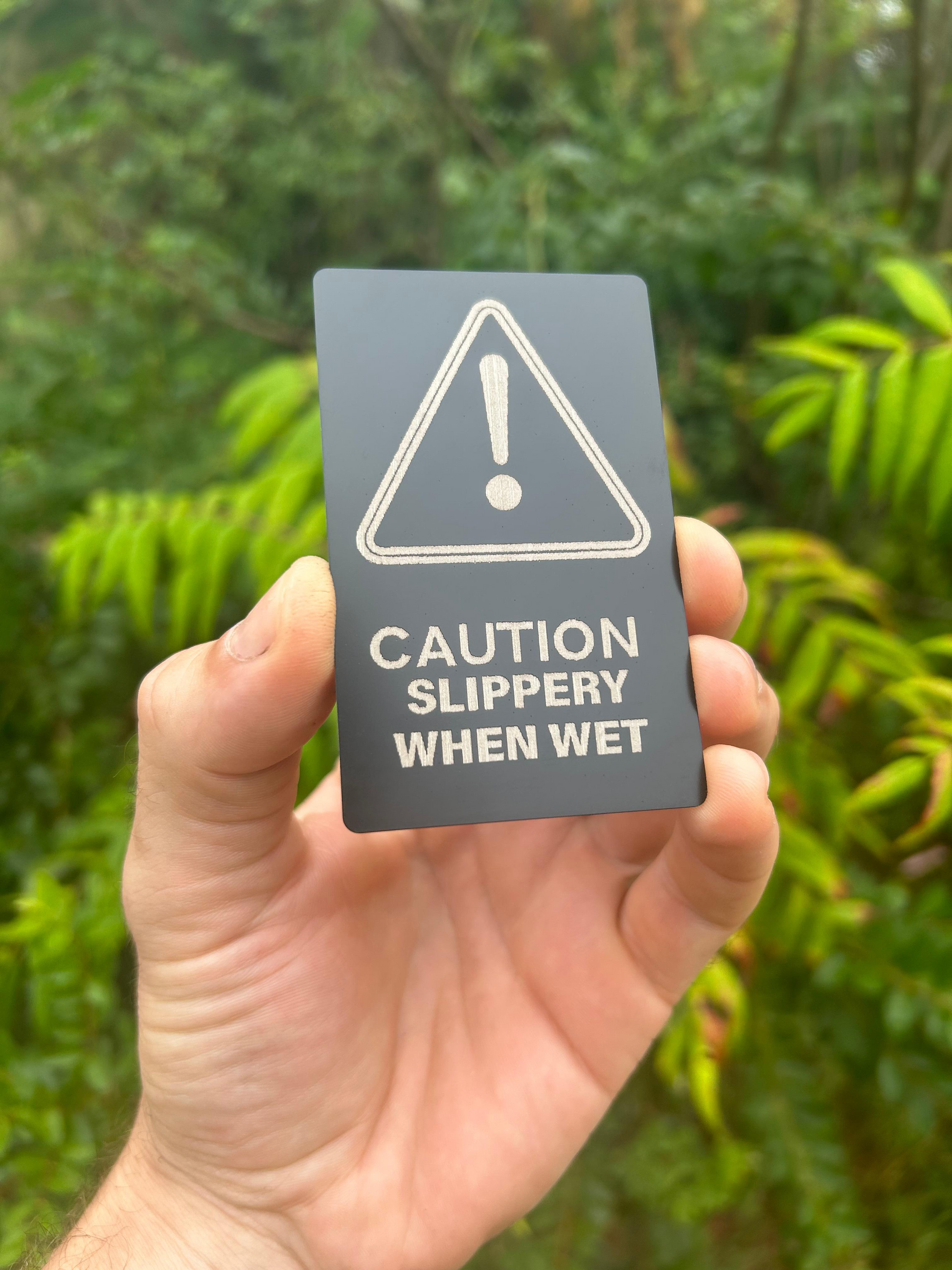 Hand holding a custom laser engraved metal black/silver anodized aluminum 'Caution: Slippery When Wet' sign with a forest background