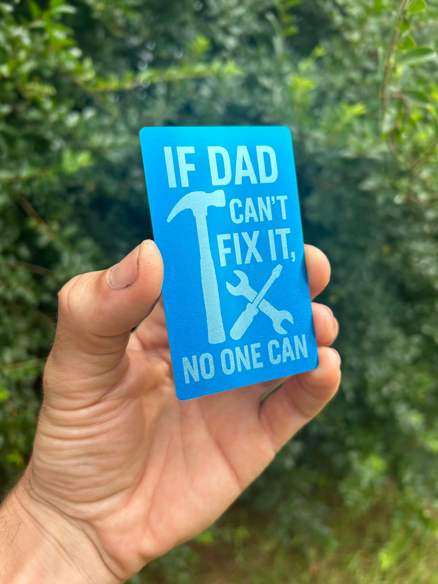 Hand holding a metal blue keepsake card with dad text against a green leafy background