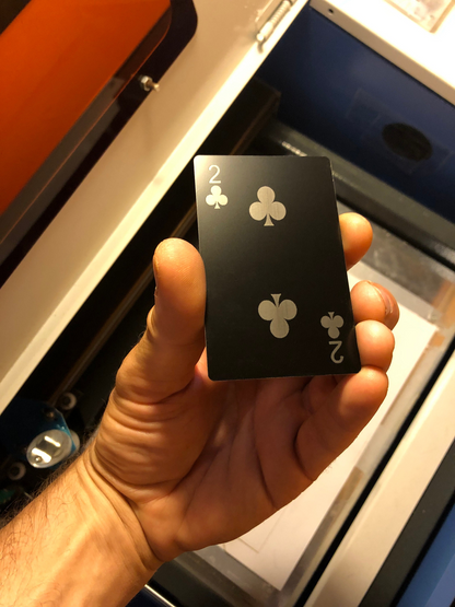 Hand holding a matte black anodized aluminum two of clubs playing card with a blue background