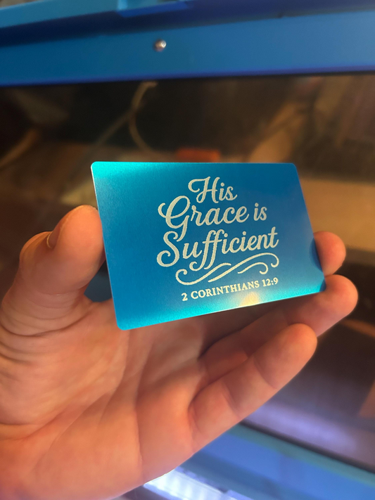Hand holding a blue and silver metal gift card with 'His Grace is Sufficient' text