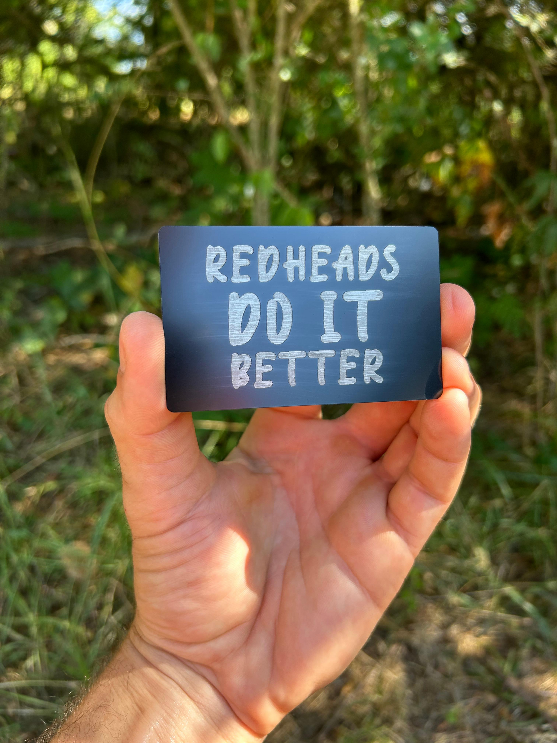 Hand holding a black card with 'Redheads Do It Better' text against a natural background
