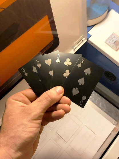Hand holding a deck of metal playing cards with Three of diamonds, Three of hearts, 3 of clubs, 3 of spades laser engraved in anodized aluminum in front of  a blurred background