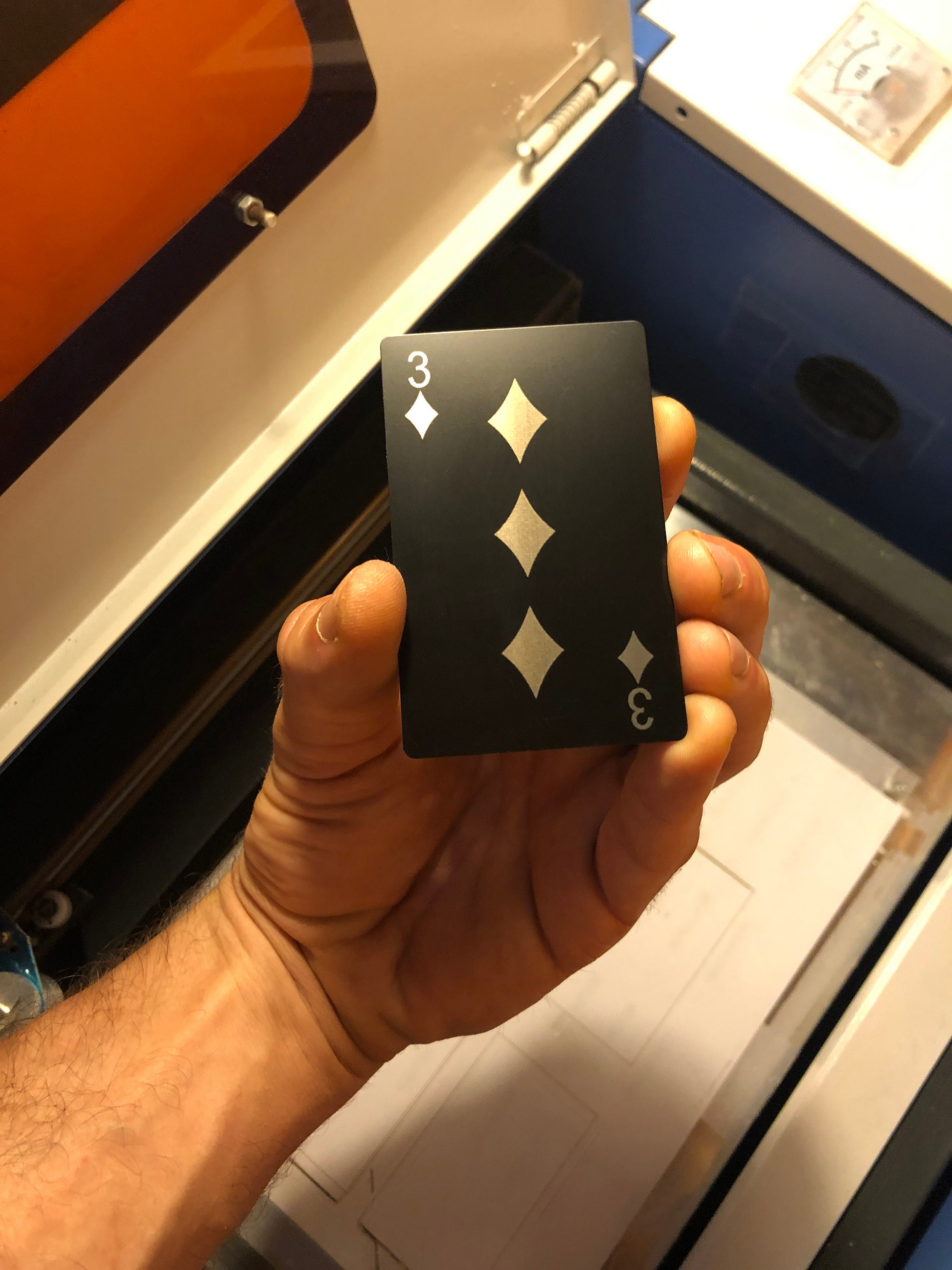 Hand holding metal gaming gift with three of diamonds laser engraved in anodized aluminum in Black/Silver in front of a laser engraving cnc machine