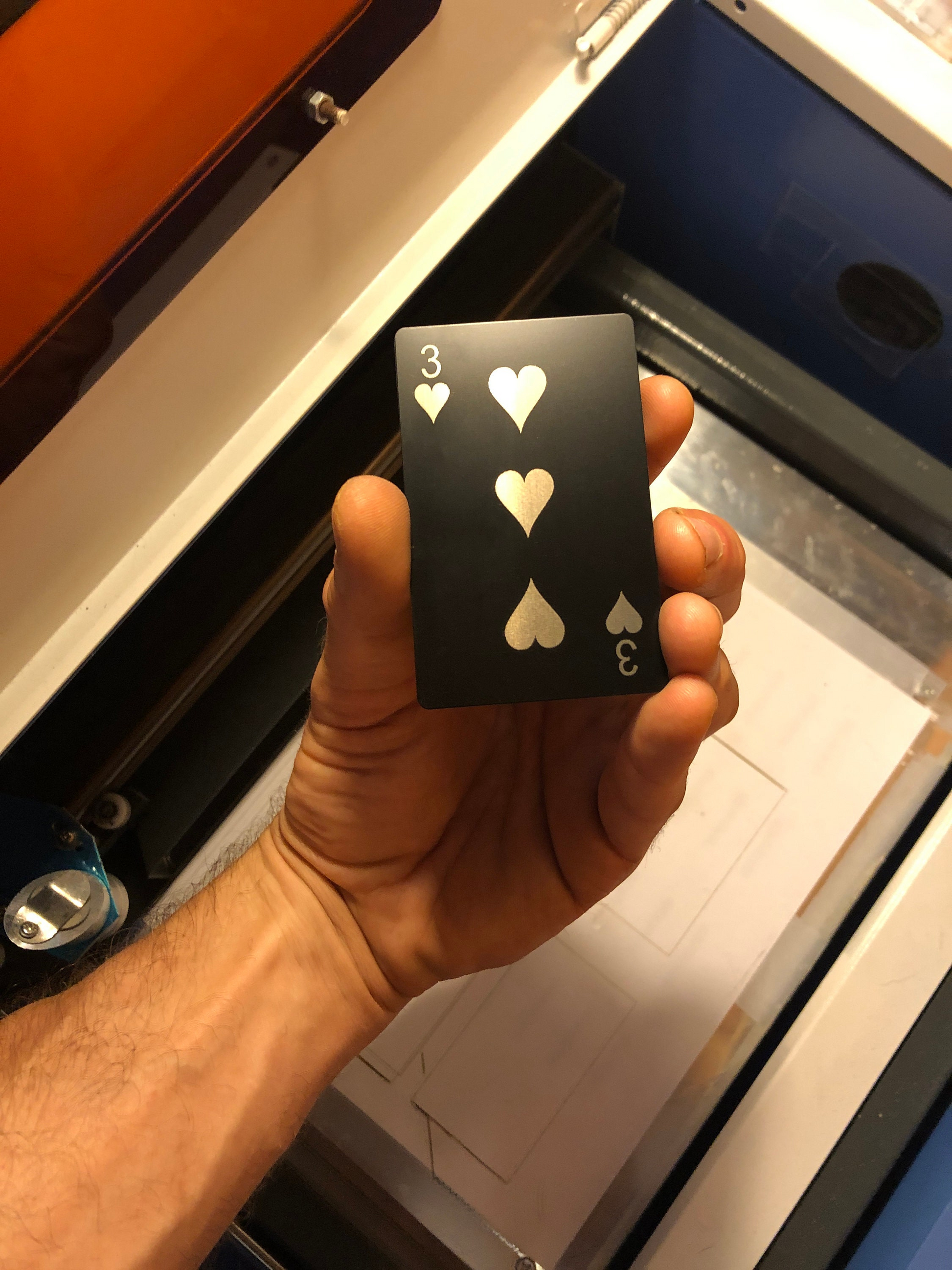 Hand holding a black metal playing card with Three of hearts laser engraved symbols in front of a machine.