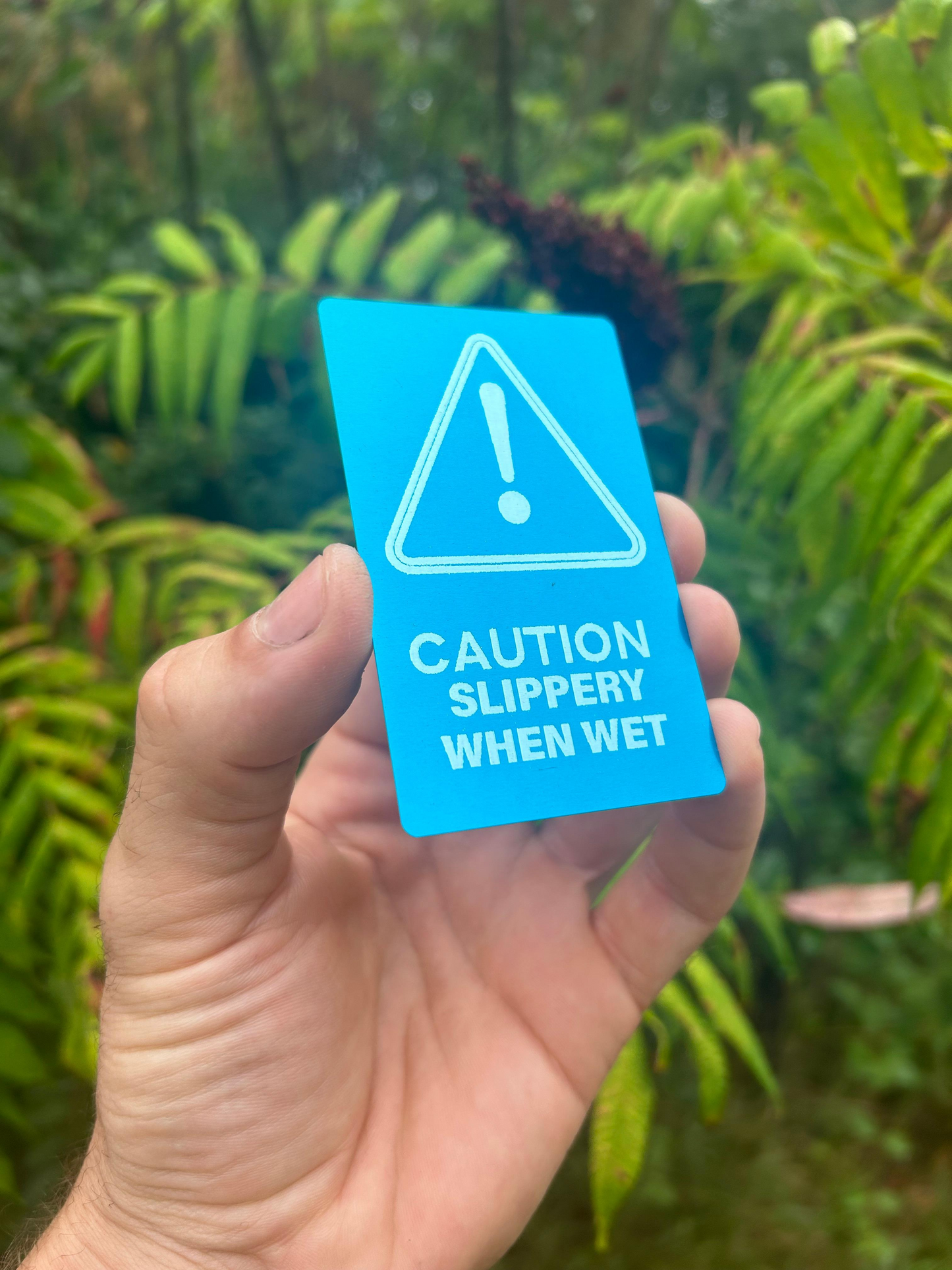 Hand holding a blue/silver metal placard with a warning symbol and text 'Caution: Slippery When Wet' against a green leafy background.
