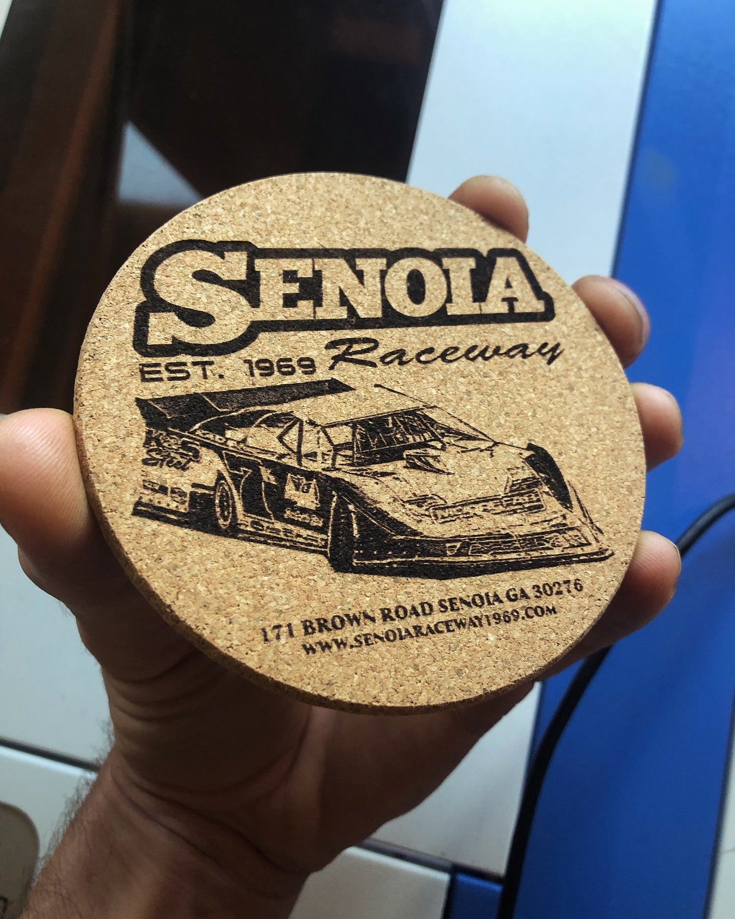 Cork coaster with Senola Raceway branding held in a hand against a blurred laser engraving machine background