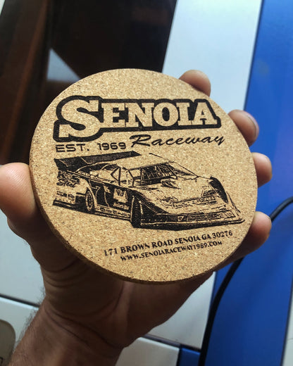Cork coaster with Senola Raceway branding held in a hand against a blurred laser engraving machine background