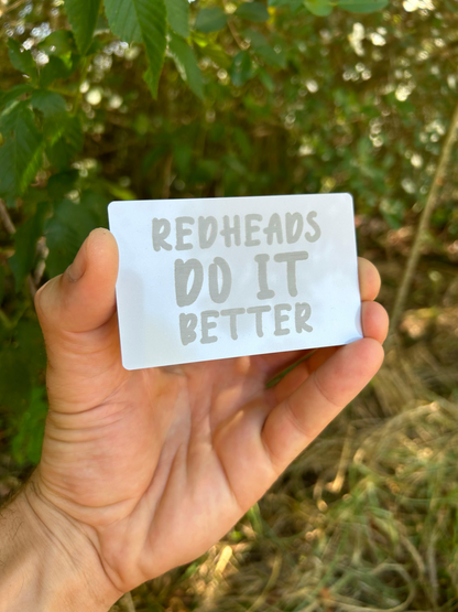 Hand holding a card with 'Redheads Do It Better' text against a natural background