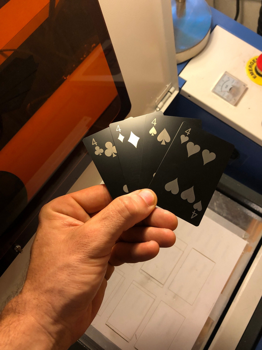 Hand holding four metal laser engraved playing cards with four of clubs, four of diamonds, four of spades, four of hearts in front of a machine and a blurred background