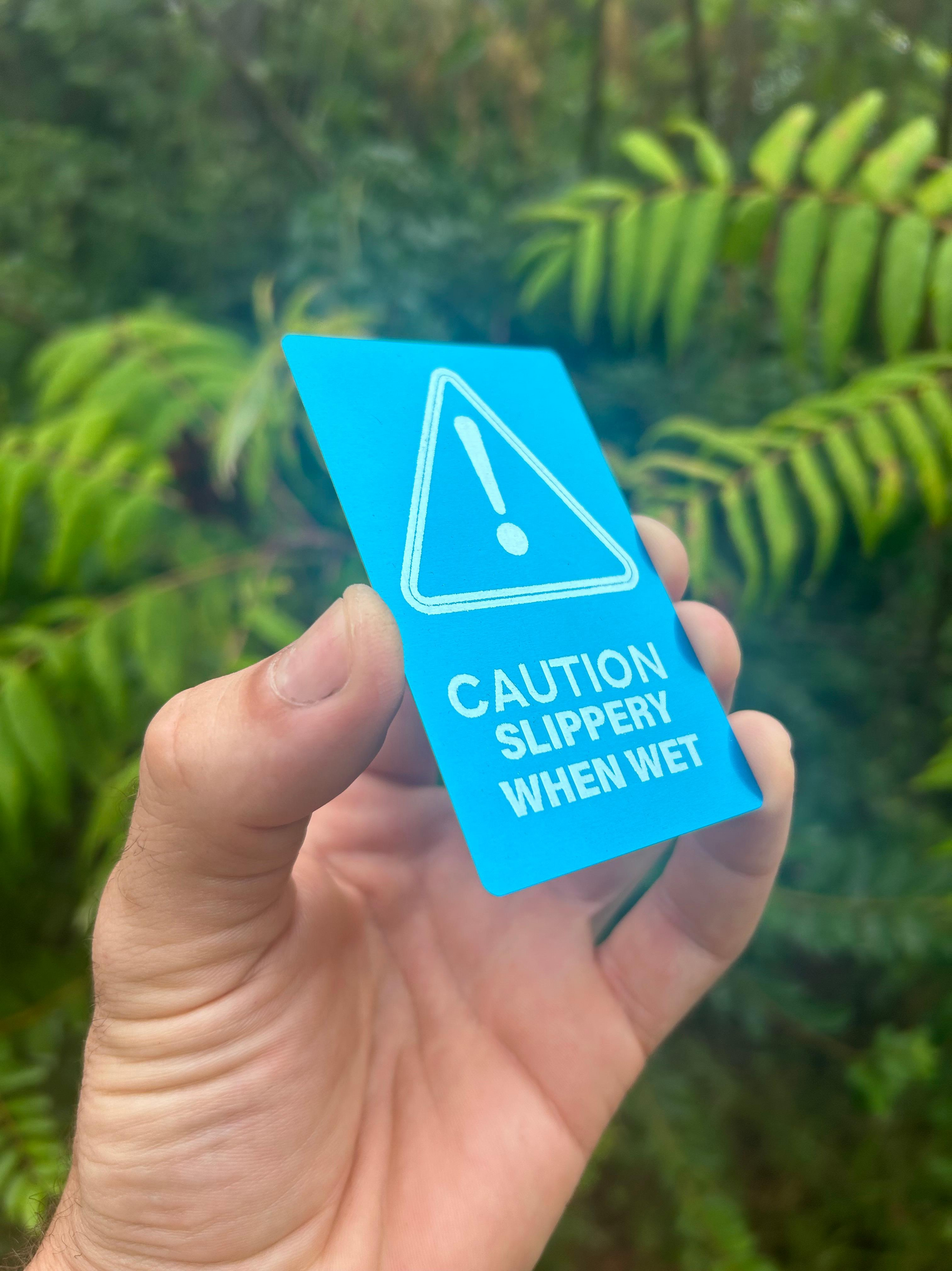 Hand holding a blue/silver anodized aluminum laser engraved 'Caution Slippery When Wet' warning signage placard against a green leafy background