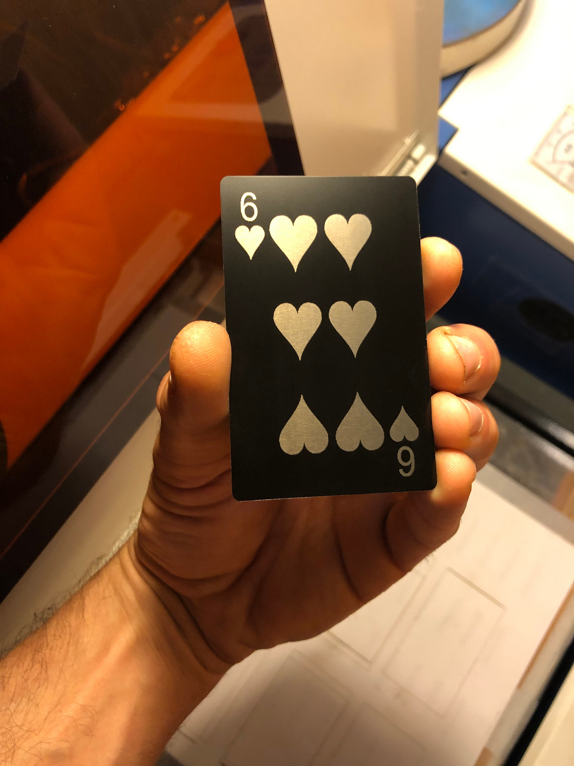 Hand holding a six of hearts metal playing card in matte black/silver in front of a blurred laser engraving machine