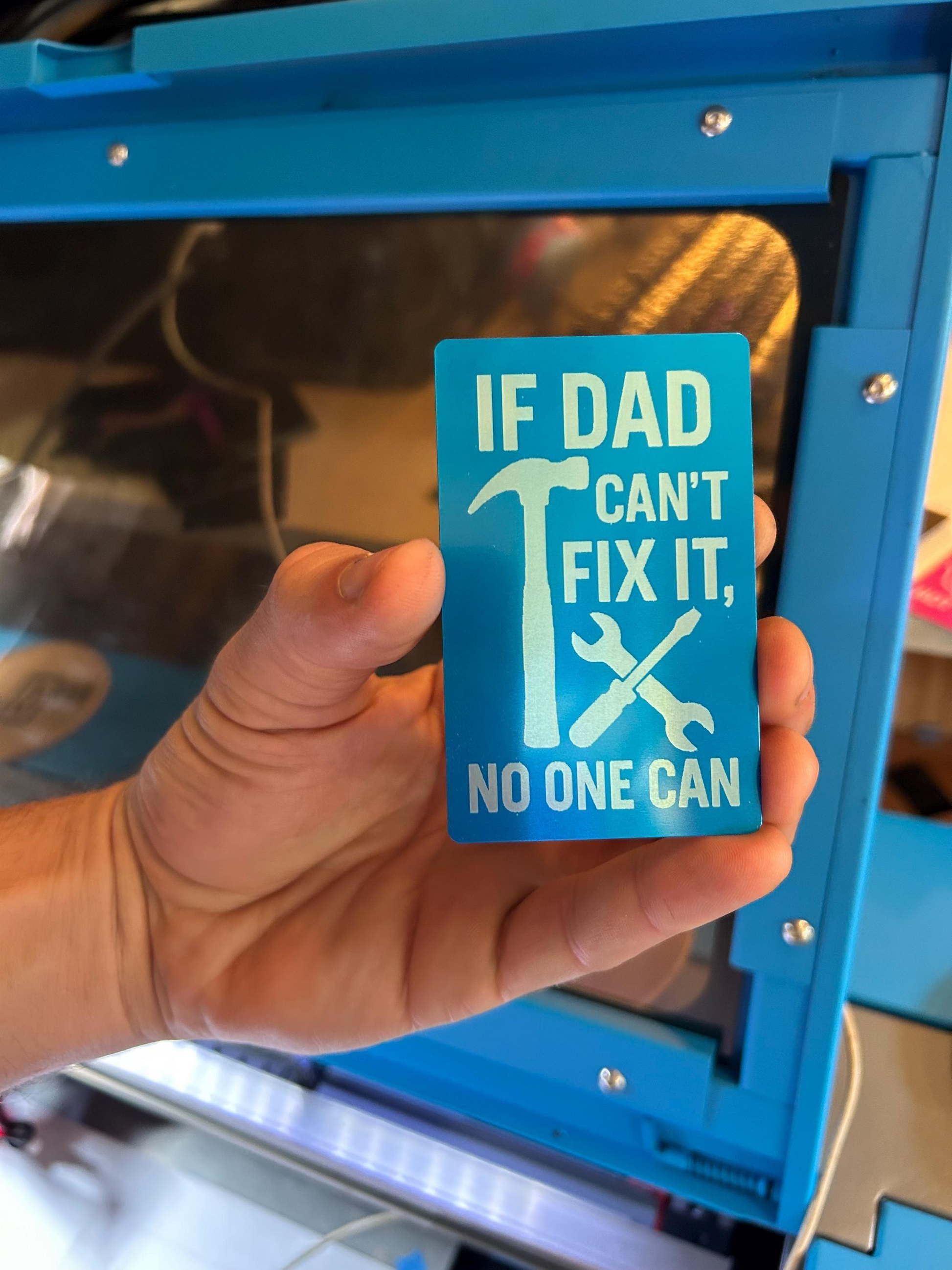 Hand holding a metal blue dad card with text and graphics in front of a blue industrial machine.