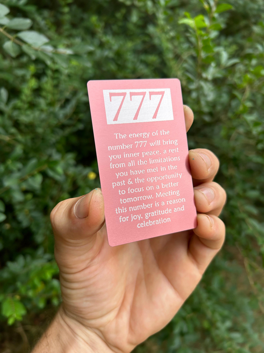 Hand holding a pink card with '777' and spiritual energy text against a green leafy background