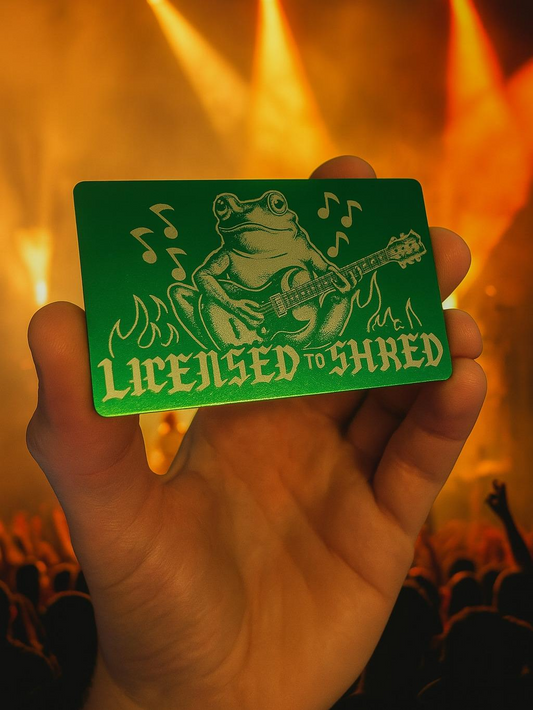 Hand holding a green card with a frog playing guitar and 'Licensed to Shred' text against a blurred concert background.