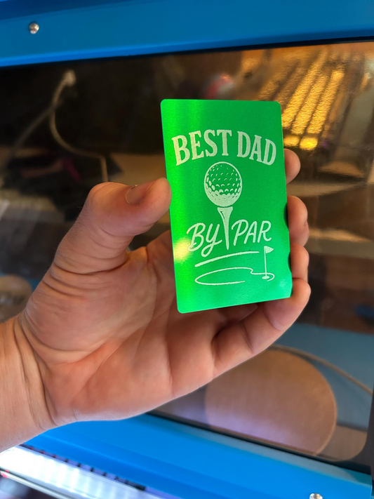 Hand holding a green/silver metal card with 'Best Dad By Par' text and golf ball design.