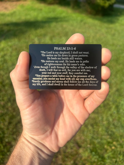 Hand holding a black metal laser engraved card with Psalm 23:1-6 text against a grassy background