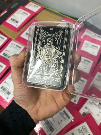 Hand holding a deck of metal tarot cards with 'Justice' in a clear plastic mailer case against a background of similar cards.