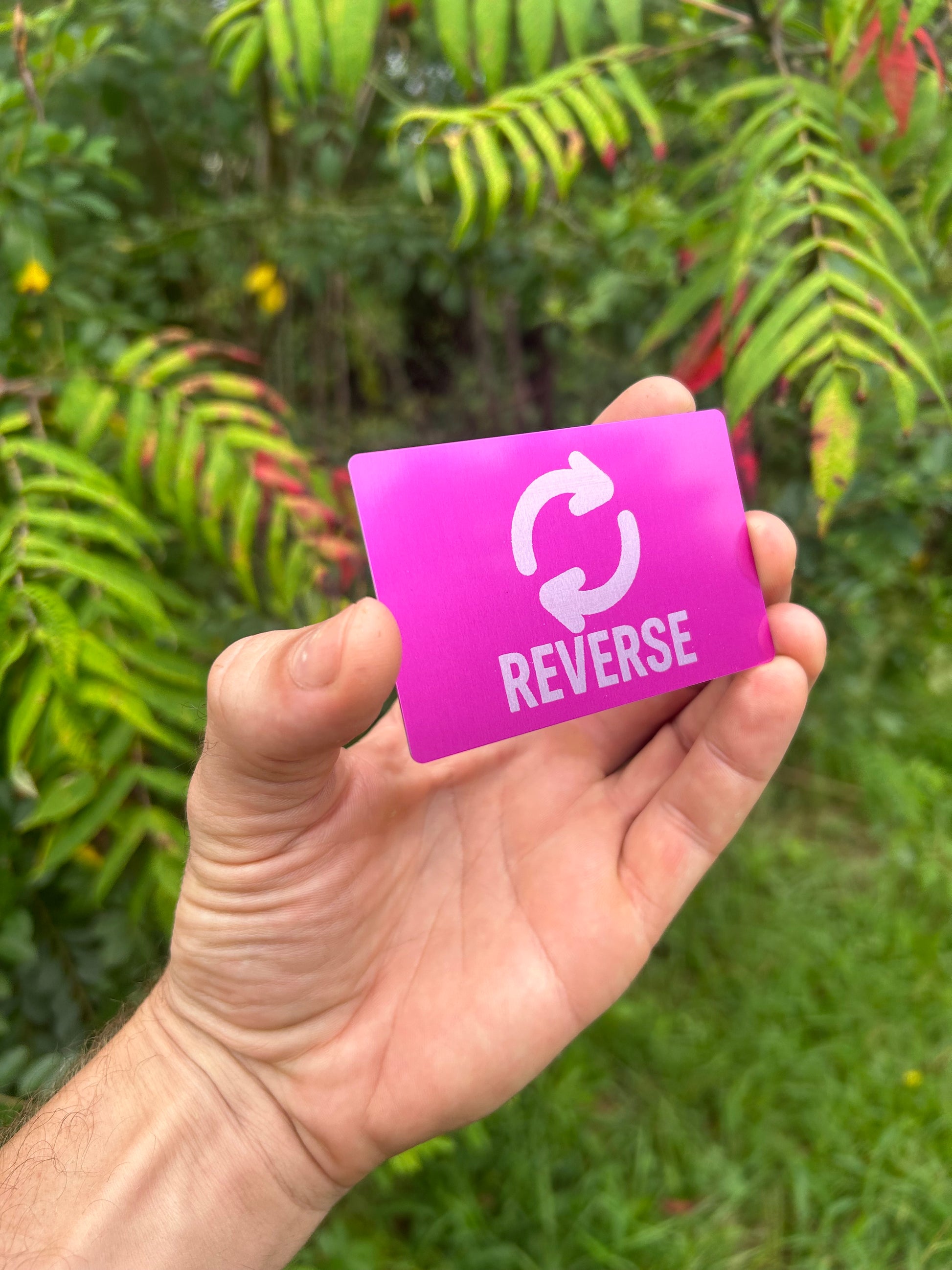 Hand holding a purple/silver metal card with 'REVERSE' text and symbol laser engraved, against a green leafy background