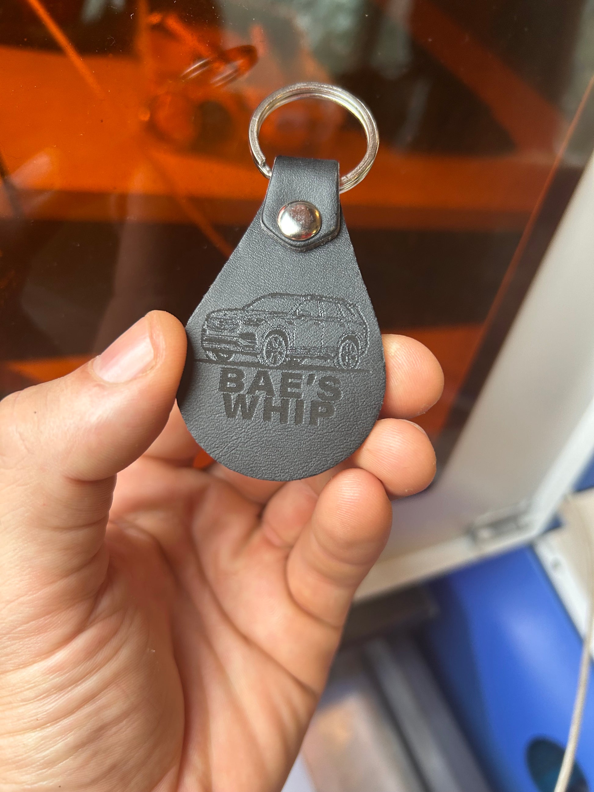Hand holding a black leather keychain with 'Bae's Whip' branding.