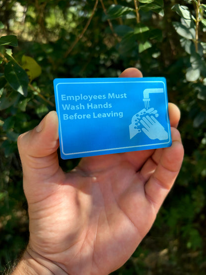 Hand holding a blue sign with text and a handwashing icon against a green leafy background