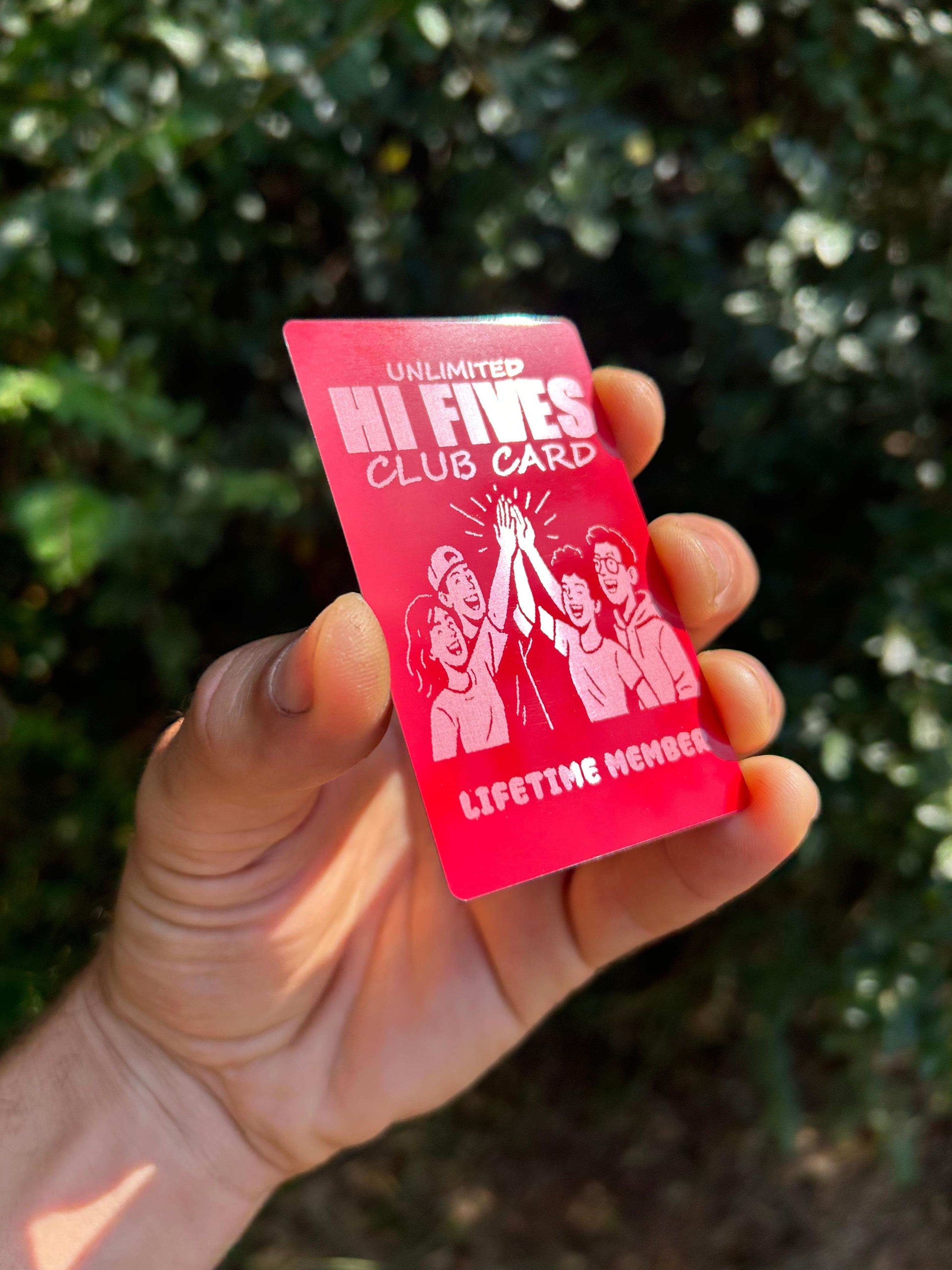 Hand holding a metal red/silver laser engraved 'Unlimited Hi Fives Club' card with an outdoor background