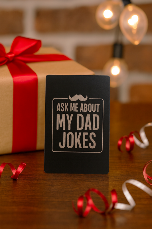 Gift with red ribbon and 'Ask me about my dad jokes' metal novelty Father's Day card on a wooden surface.