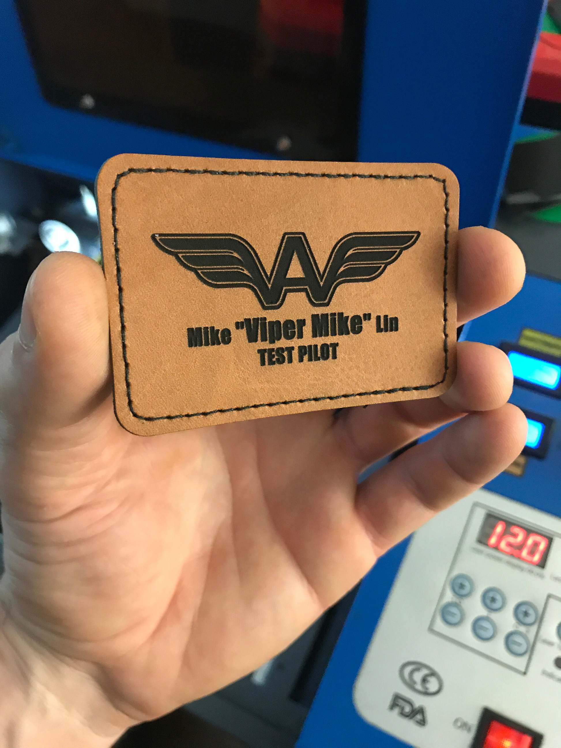 Brown leather patch with 'Mike Viper Mike' and 'TEST PILOT' text held by a hand against a blue machine background.