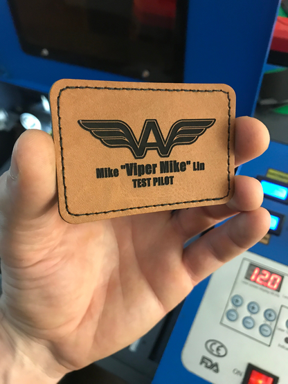 Brown leather patch with 'Mike Viper Mike' and 'TEST PILOT' text held by a hand against a blue machine background.