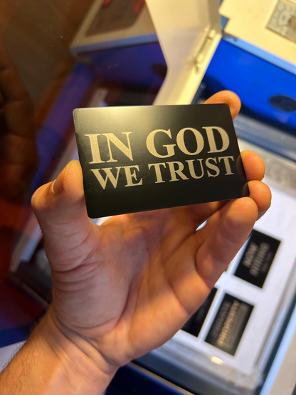 Hand holding a metal business card with 'IN GOD WE TRUST' text against a blurred background