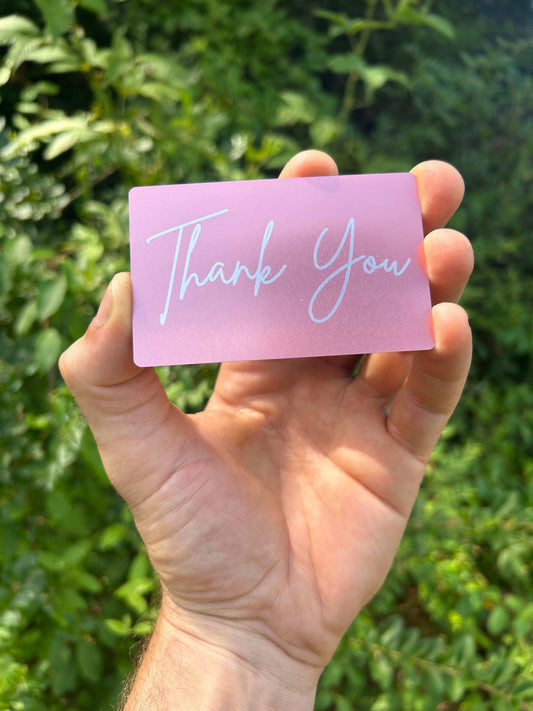 Hand holding a pink metal card with 'Thank You' text against a green leafy background
