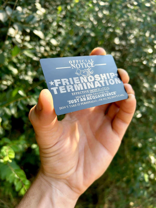 Hand holding a funny novelty card with 'Official Notice: Friendship Termination' text against a green leafy background