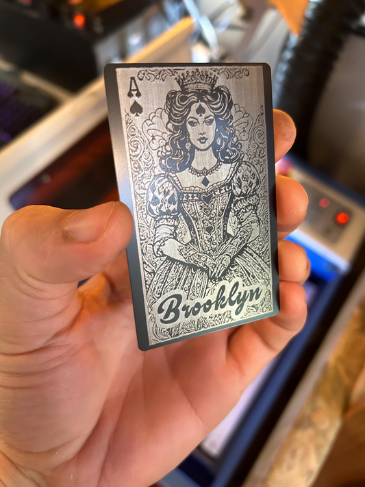 Close-up of hand displaying laser engraved metal Ace of Spades card with Lady Luck artwork against blurred background