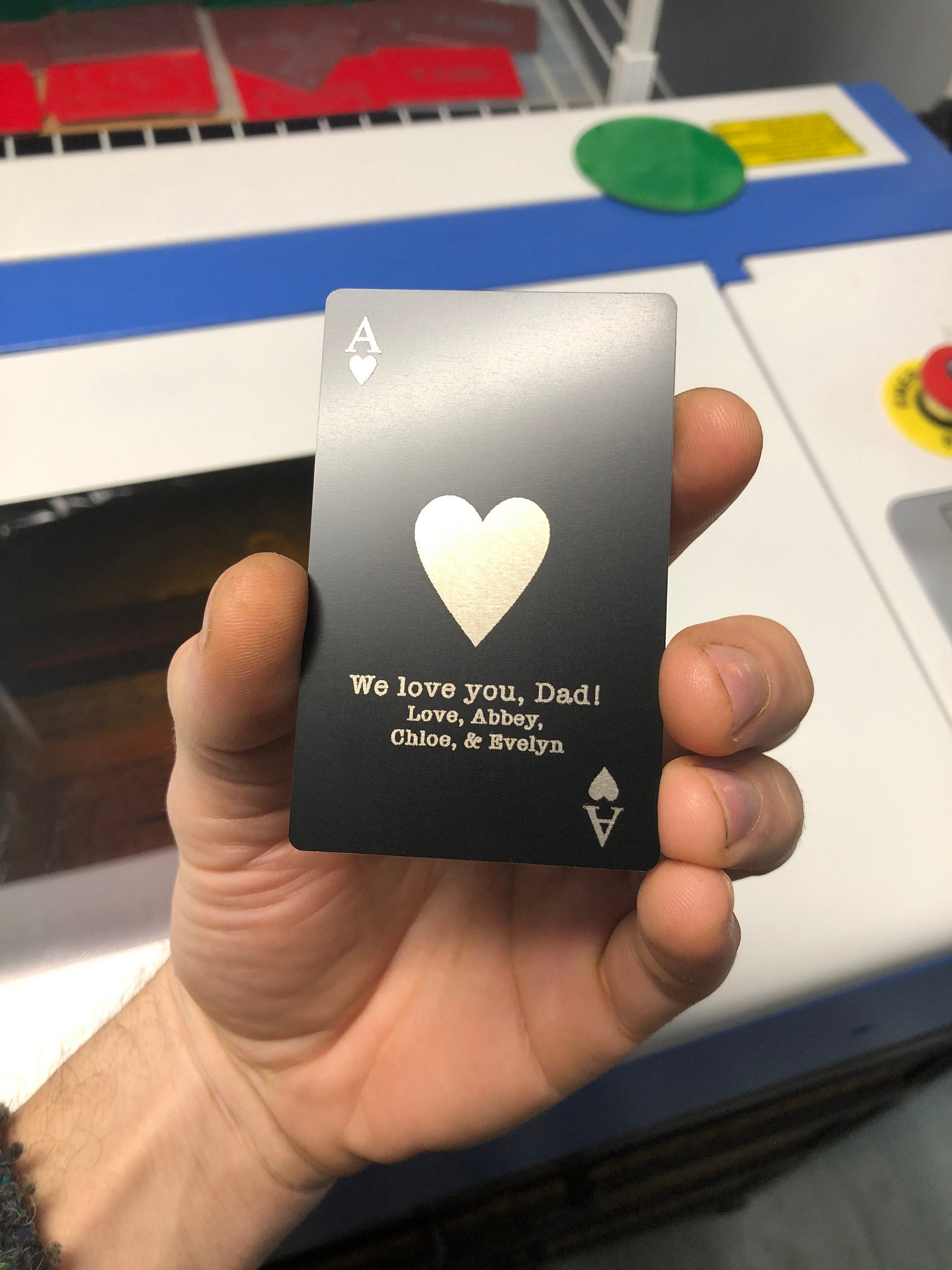 Hand holding a black Ace of Hearts playing card with a silver heart and personalized text in front of a machine.
