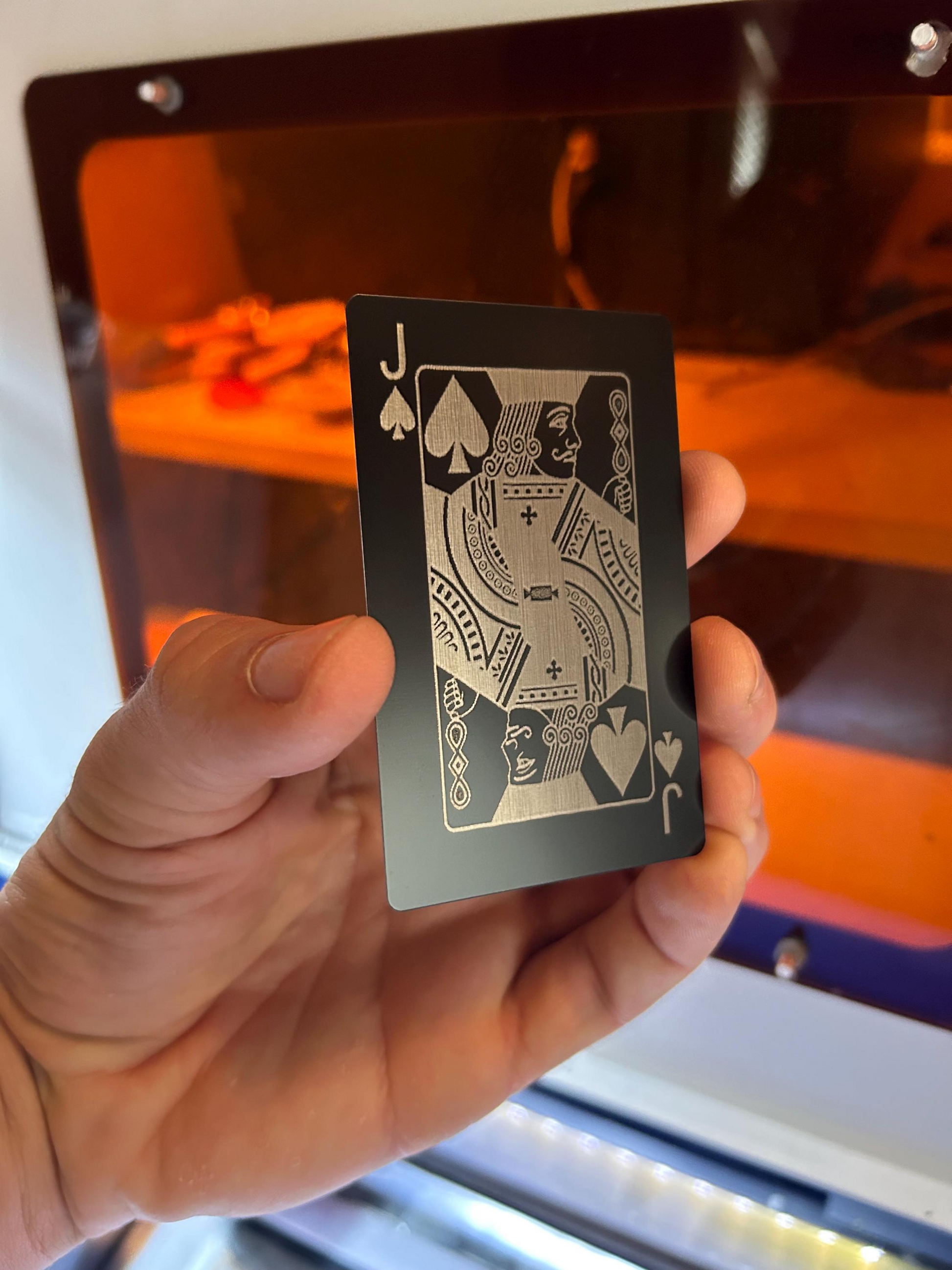 Hand holding a black and silver metal Jack of Spades poker card, laser engraved in anodized aluminum in front of a machine background