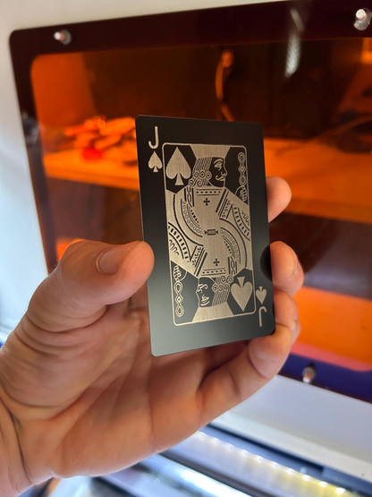 Hand holding a black and silver metal Jack of Spades poker card, laser engraved in anodized aluminum in front of a machine background