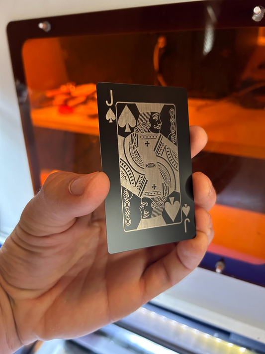 Hand holding a black and silver metal Jack of Spades poker card, laser engraved in anodized aluminum in front of a machine background
