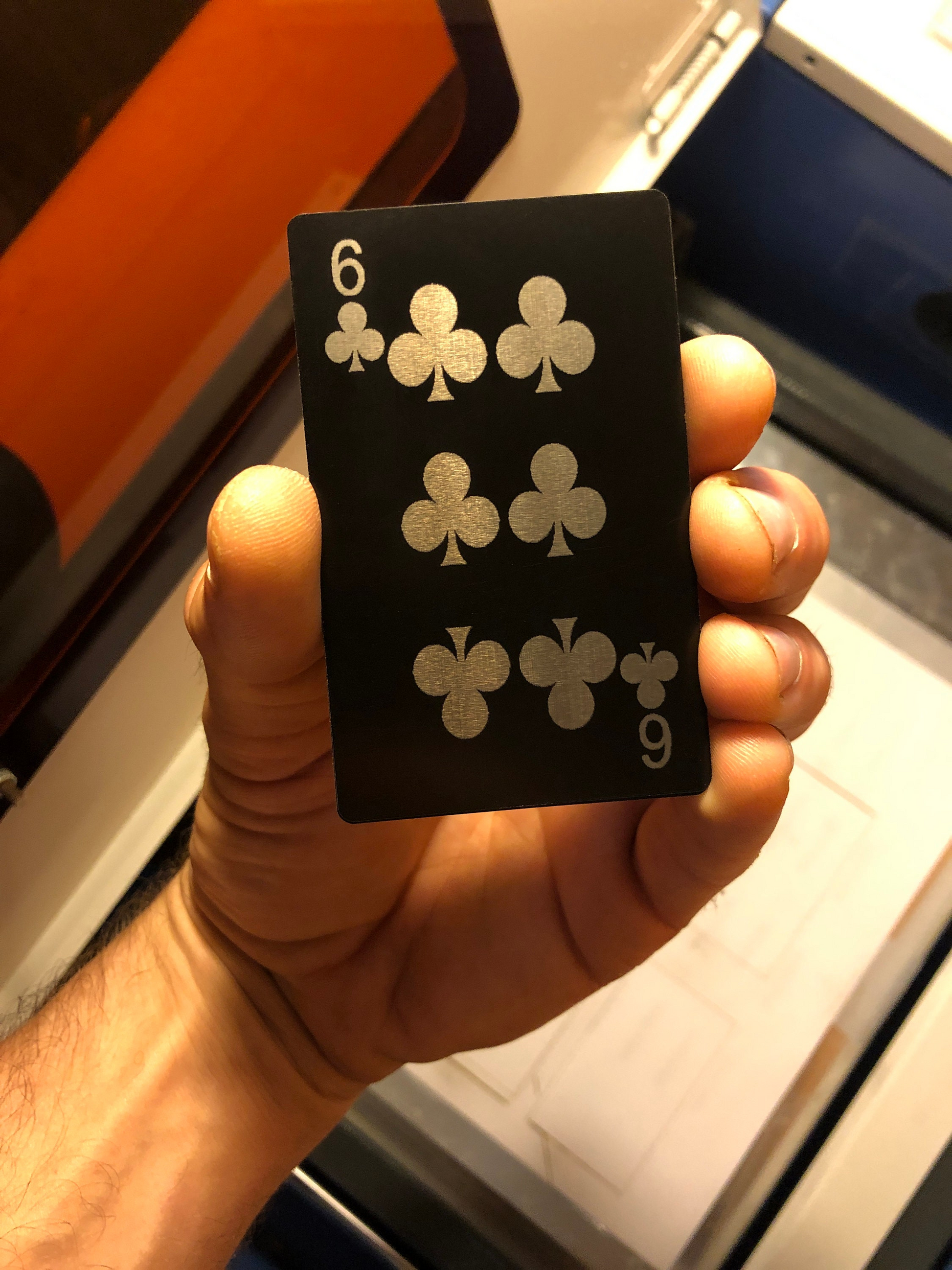 Hand holding matte black/silver anodized aluminum laser engraved poker card with Six of Clubs design in front of a blurred machine background