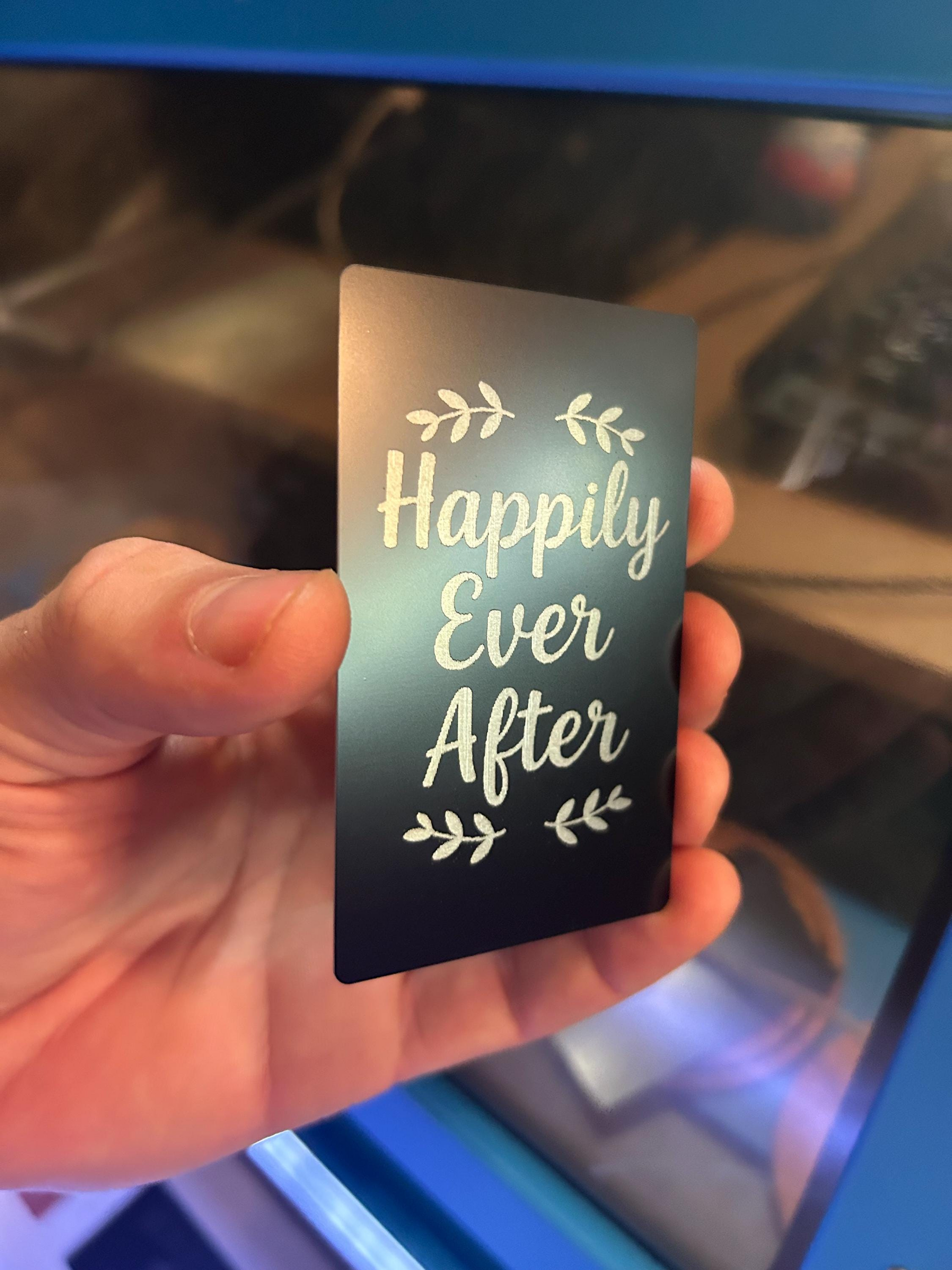Person holding a laser engraved anniversary gift card in black and silver metal with “Happily Ever After” design