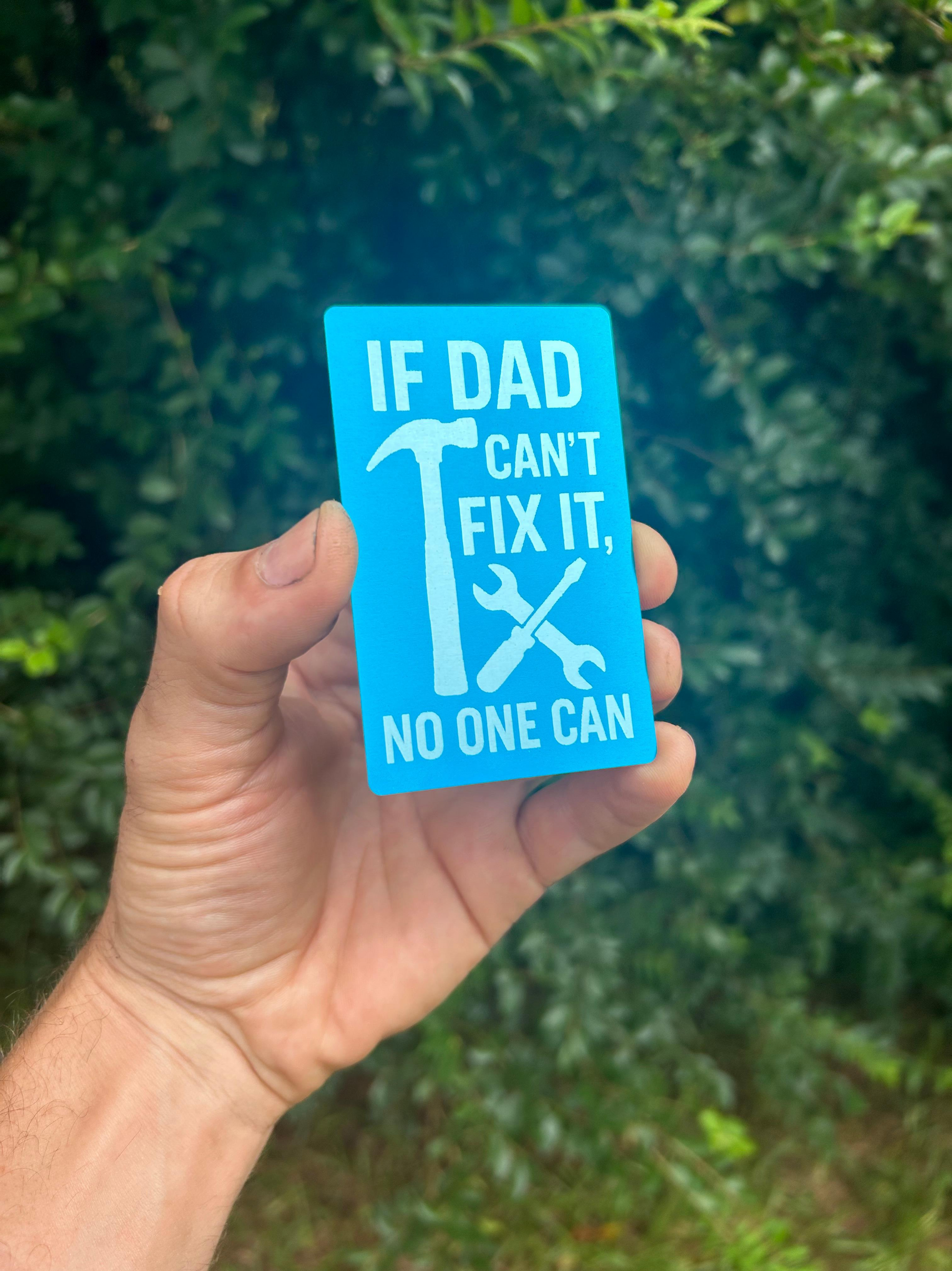 Hand holding a blue metal DAD card with text and tools graphic against a blurred green background
