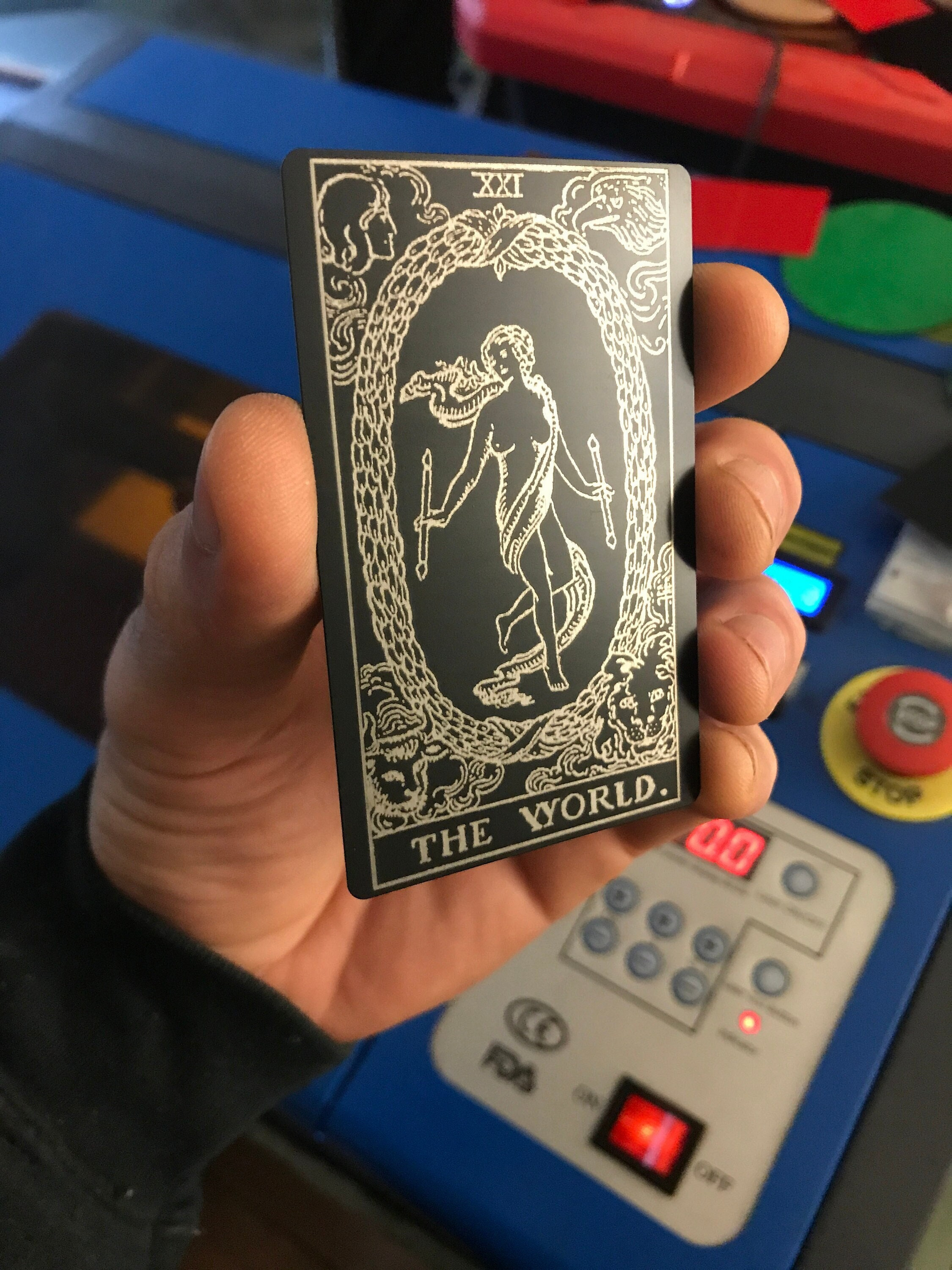 Hand holding a laser engraved metal tarot card titled 'The World' in front of a control panel.