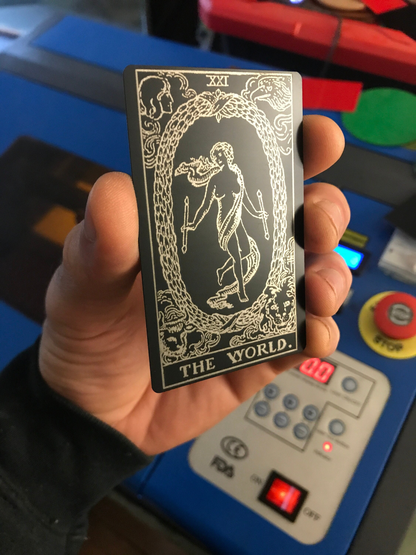 Hand holding a laser engraved metal tarot card titled 'The World' in front of a control panel.