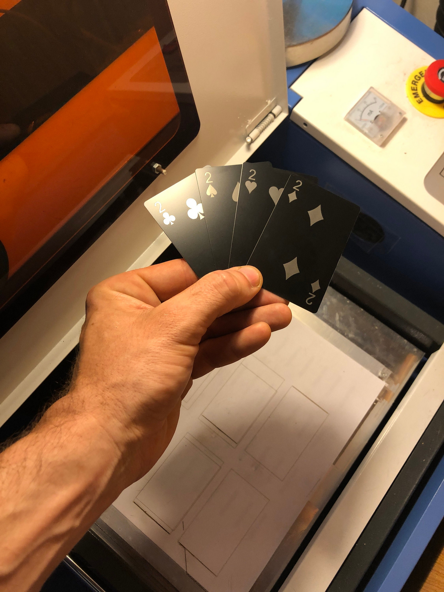 Hand holding a two of clubs, two of spades, 2 of hearts, 2 of diamonds laser engraved metal poker cards in front of a production laser engraving machine